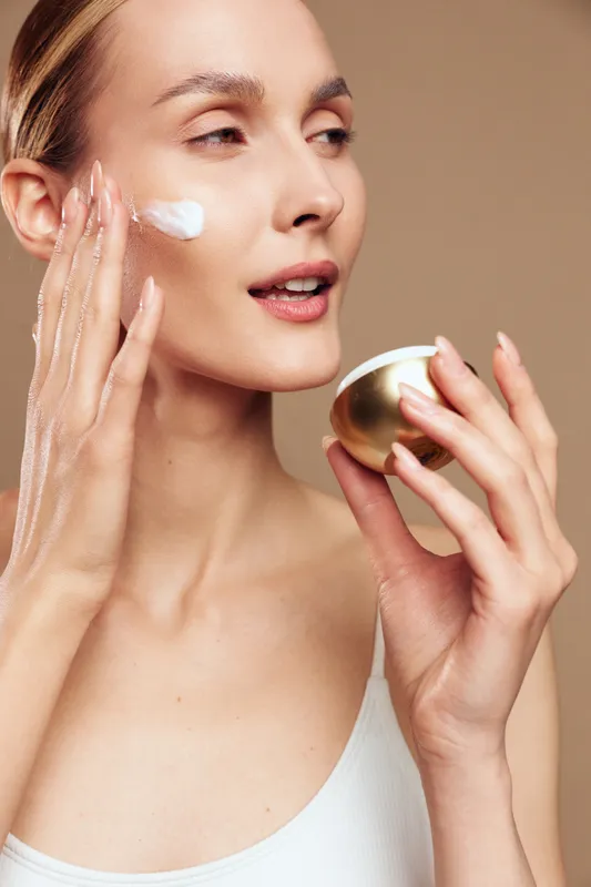 Young woman with clear skin applies moisturizing cream on her face holding cosmetic jar, skincare routine concept, natural beauty care isolated on beige background.