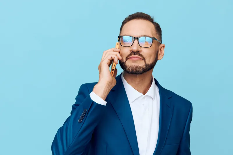 A professional man in a blue suit speaks on a mobile phone against a solid colored backdrop, portraying confidence, focus, and determination in a business moment.
