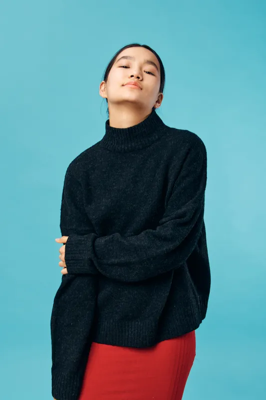 Young woman in black sweater and red skirt posing with folded arms on blue background. Casual outfit, confident expression, indoor studio portrait with minimalistic style and soft lighting.