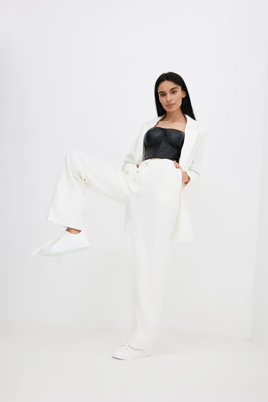 Young woman posing confidently in white suit and black top, showcasing modern fashion style with minimalistic white background and casual sneakers.