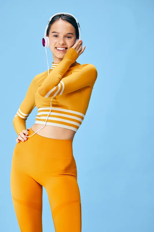 Young woman fitness model wearing bright orange activewear and white headphones, smiling confidently while posing with hand on hip against a clean blue studio background, energetic sporty concept.