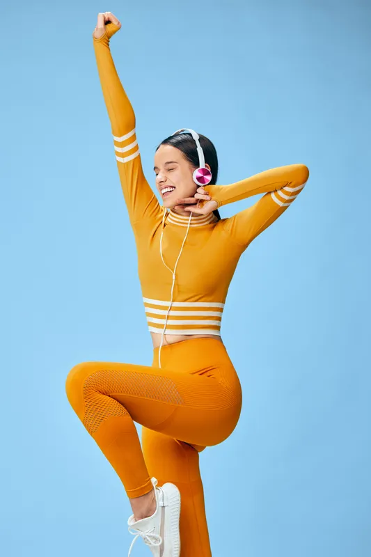 Young woman fitness model in orange athleisure posing mid-stretch with one knee raised, wearing headphones and sneakers against solid blue studio background and upbeat energy.