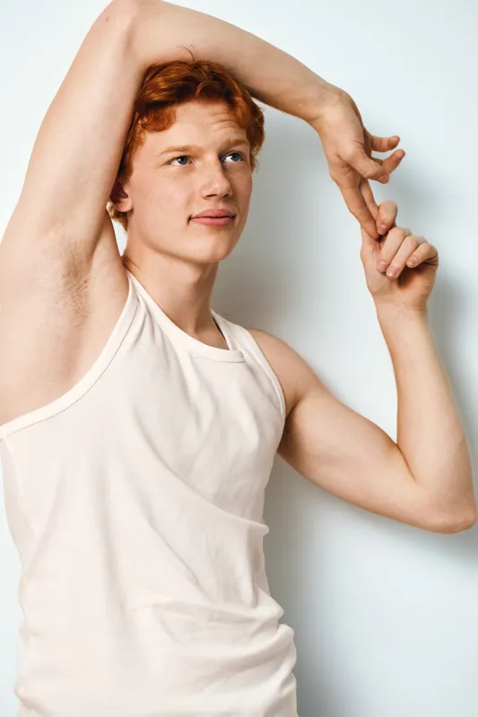 Young man with red hair stretching arms above head wearing white sleeveless shirt standing against white wall for fitness relaxation and morning routine at home or studio.