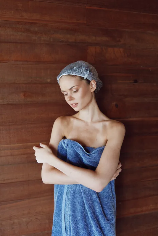 young woman wrapped in blue towel with shower cap standing against wooden wall, natural light, indoor bathroom, fresh skincare moment