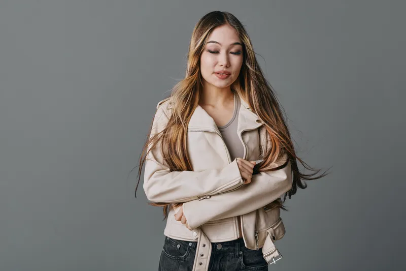 Young woman with long hair wearing light jacket standing with crossed arms and closed eyes isolated on gray background in studio portrait of confident fashion model.