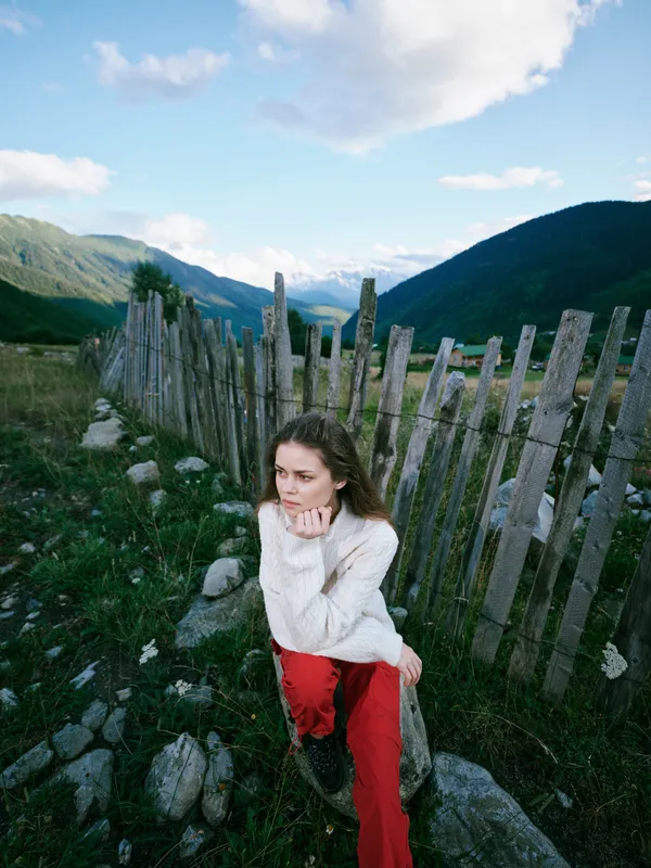 Woman sits beside a weathered fence in a mountainous countryside landscape, thoughtful expression, casual white sweater and red pants, capturing a serene outdoor moment