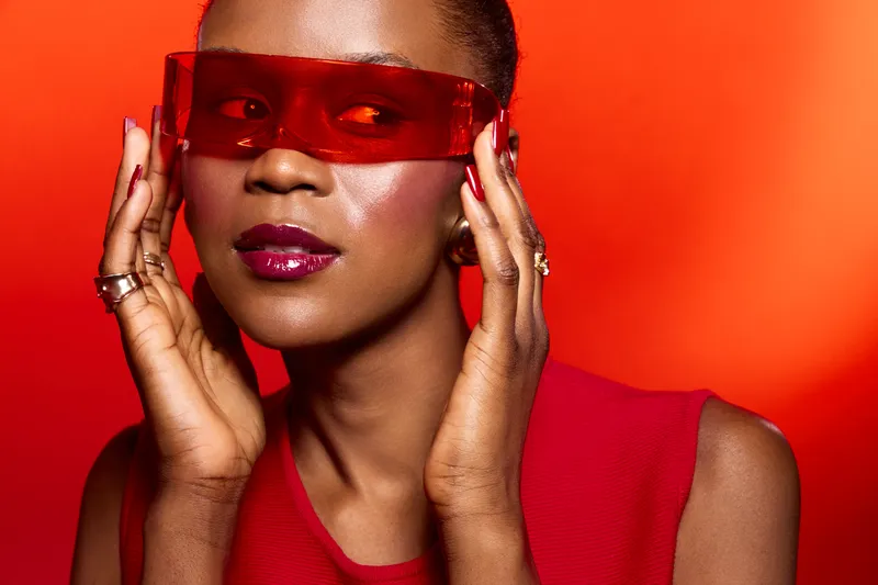 Confident African woman with dark skin wearing red sleeveless top and red translucent sunglasses poses against bright red background. Fashion and beauty concept with bold colors and style.