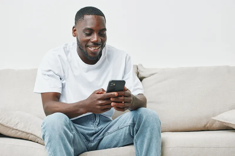 Happy African American man using smartphone on sofa in casual clothing, enjoying leisure time at home, smiling and relaxed, showcasing modern lifestyle