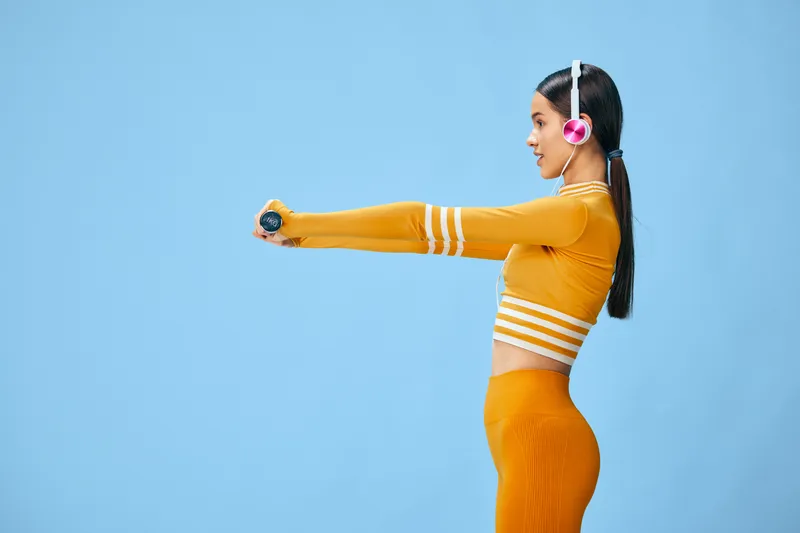 Female fitness athlete in orange sportswear extends arms forward in a studio setting with headphones, showcasing balance, movement and athletic energy against a blue backdrop.