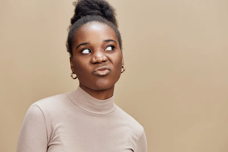 Thoughtful young African woman with an expressive look, wearing a beige turtleneck, against a warm, neutral background, reflecting emotions of contemplation and curiosity