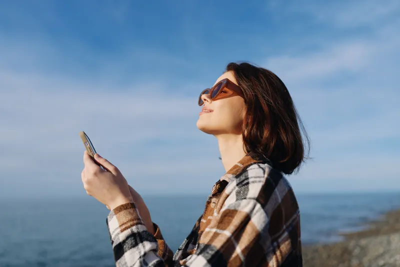 Woman in a plaid shirt and sunglasses using a smartphone by the sea, enjoying sunlight and breeze with the ocean horizon in the background