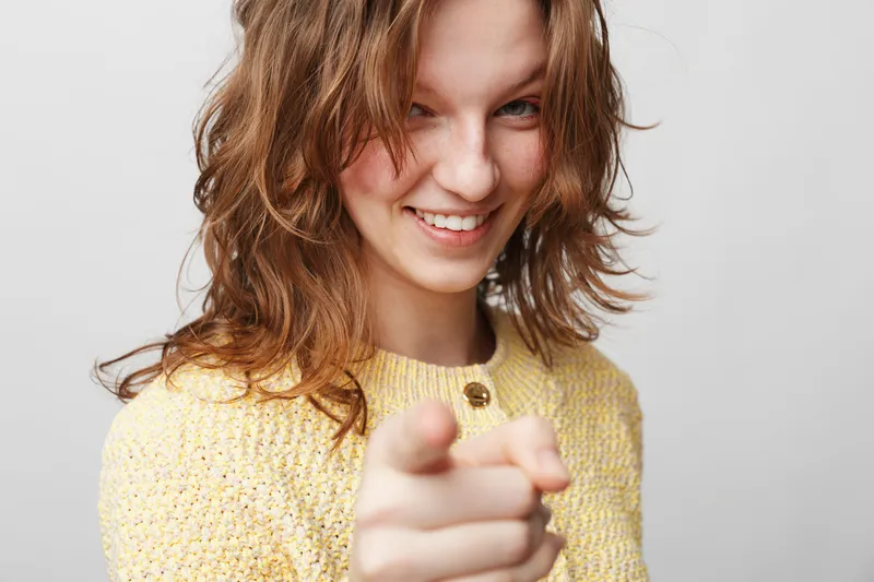 Smiling young woman with wavy brown hair wearing yellow sweater points finger at camera. Studio portrait on grey background. Concept of confidence, friendliness, and positive interaction.