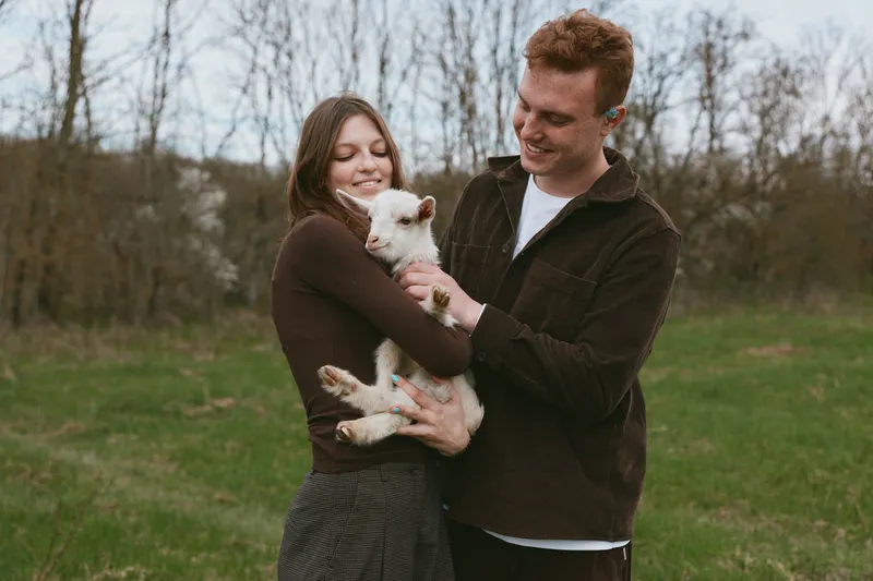 Smiling couple outdoors cradles a joyful goat kid, highlighting care, affection, and a warm lifestyle moment. Natural light on a green field conveys gentle togetherness and happiness.