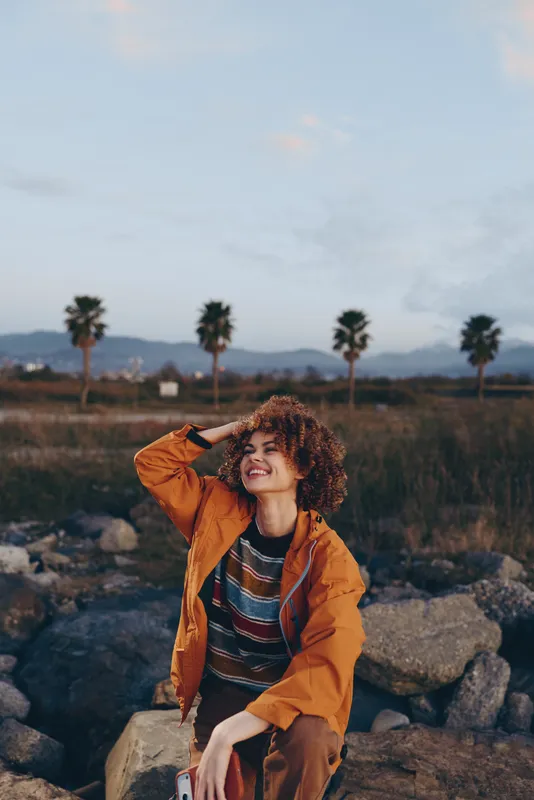 Woman lifestyle outdoors portrait, curly hair, orange jacket, smiling pose near palm trees and rocks, nature scenery at sunset, relaxed travel mood, soft clouds