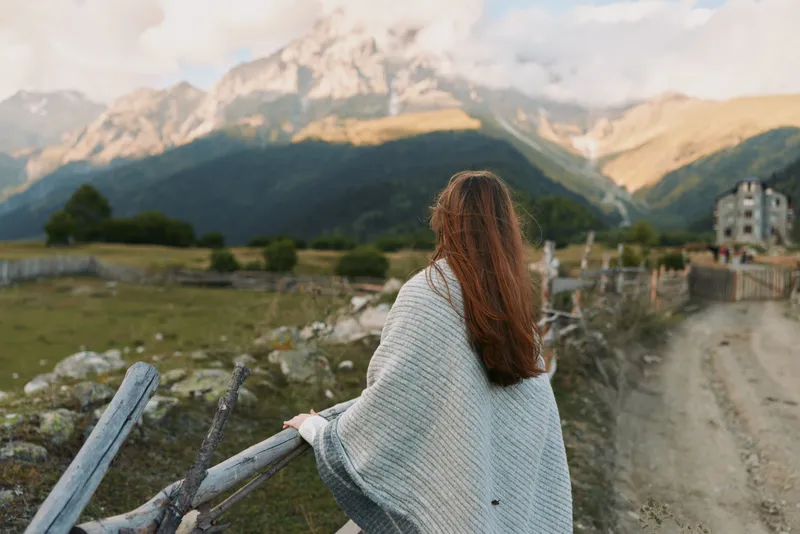 Outside scene with a woman seen from behind, wearing a cozy cardigan, gazing toward distant mountains, a relaxed lifestyle moment in natural light.