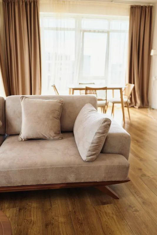 Sofa with cushion in bright living room with hardwood floor and sheer curtains, modern neutral decor, cozy seating and soft textures in a sunlit apartment interior for relaxation.