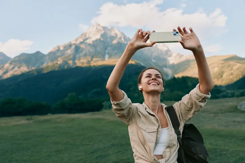 A happy woman in casual clothes takes a selfie with a smartphone in a green field with mountains in the background during a sunny day. Lifestyle and travel concept.