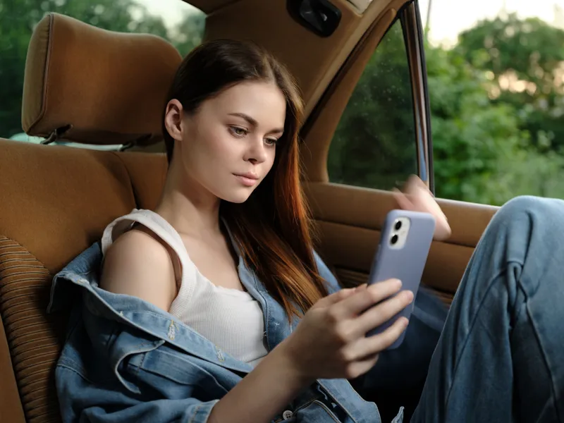 Young woman in casual denim outfit using smartphone in car, enjoying summer day, relaxation, nature background, modern lifestyle, youthful spirit.