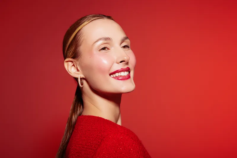 Smiling woman with red lipstick and shiny skin poses against red background. She wears red clothing and gold earrings. Concept of beauty, fashion, confidence, and vibrant style.