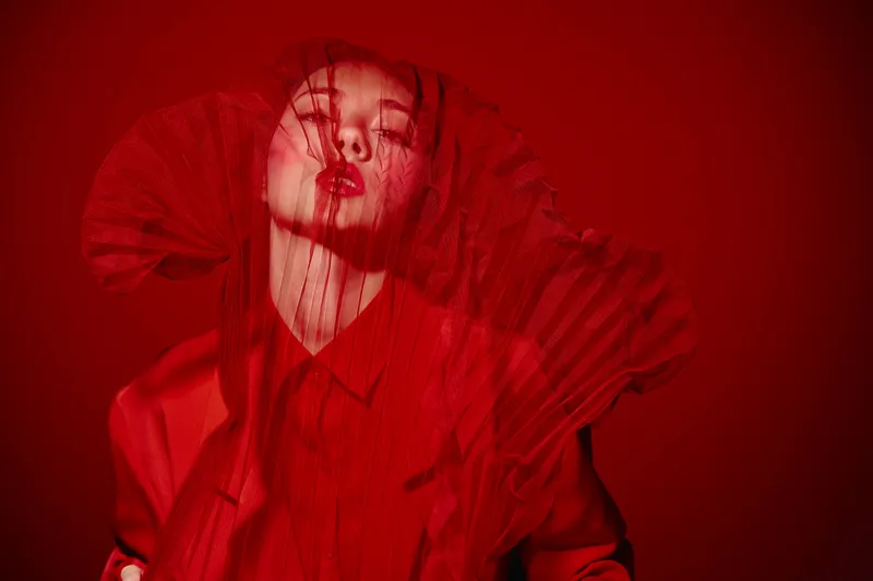 Artistic beauty portrait of a fashion model with red veiled fabric covering her face in a creative, dramatic red background studio setting. Concept of avantgarde style and editorial fashion.
