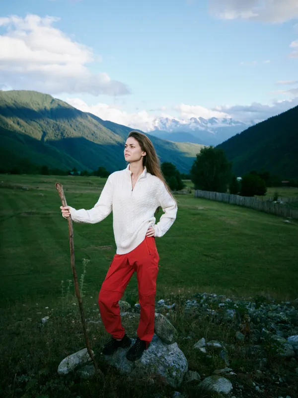 Woman standing on a rocky meadow with a hiking staff, mountains in the distance, clear sky, outdoor adventure vibe, tranquil countryside scenery and open valley