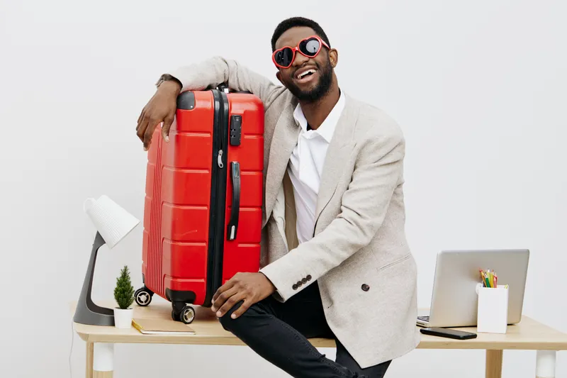Stylish cheerful man with sunglasses, dressed in light blazer and shirt, smiling happily and sitting at desk with large red suitcase, laptop, and office supplies on white background. People lifestyle 