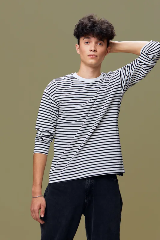 Young man with curly hair wearing a striped long sleeve shirt posing with hand behind head against brown background in casual style portrait.