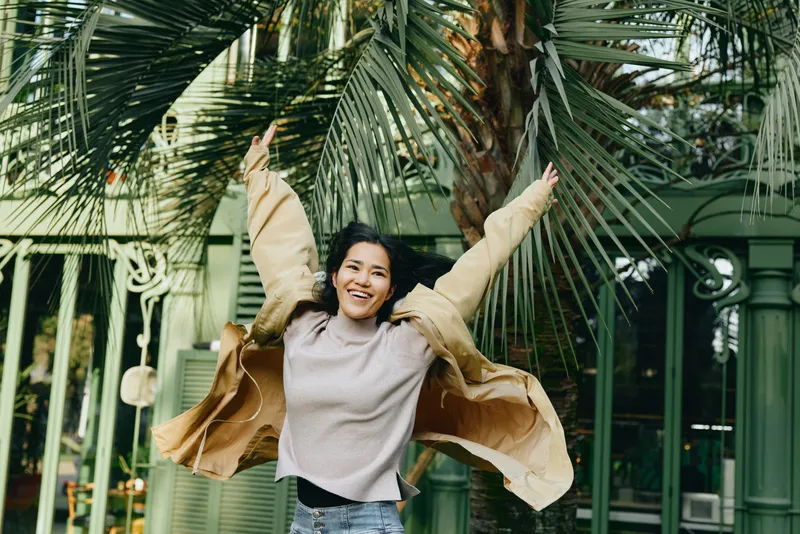 Joyful woman outdoors with flowing bags like wings, in a leafy urban setting, celebrating fashion and movement, casual chic, wind catching fabric, sunny street scene, vibrant energy