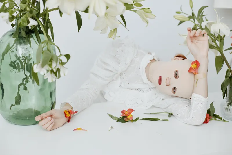Surreal portrait with a doll like model surrounded by flowers, evoking emotions of serenity and whimsy, showcasing delicate white and vibrant colors