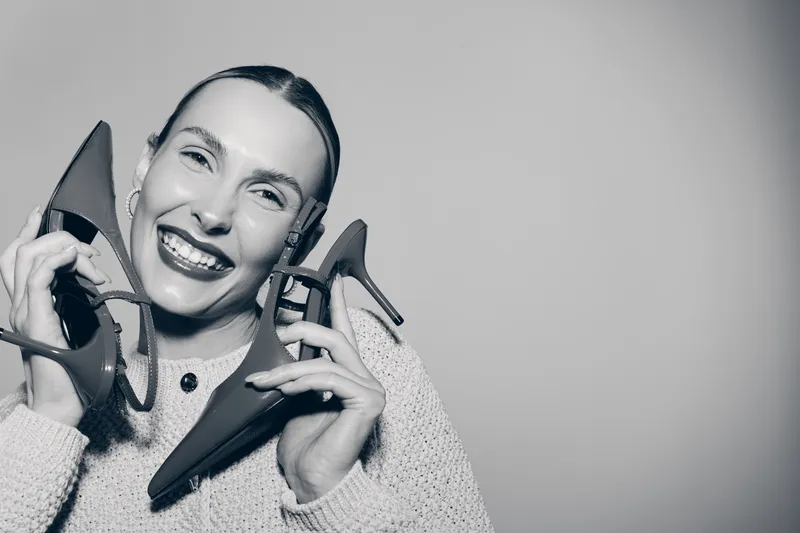 Happy Caucasian woman with fair skin smiling broadly holds two high heel shoes near face wearing knitted sweater in grey studio background. Concept of fashion and joy.