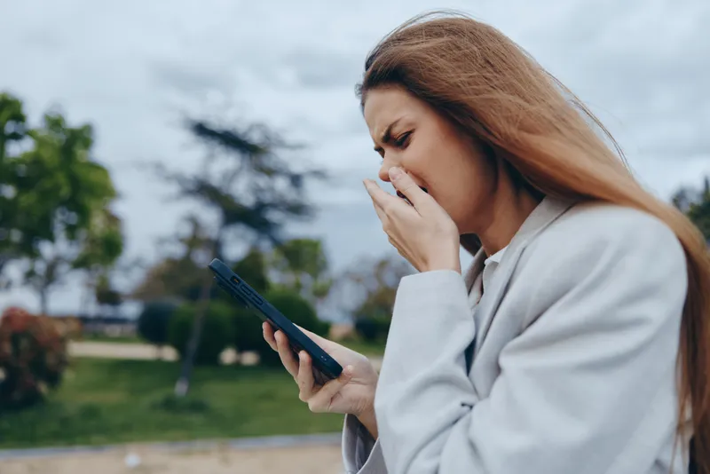 Young woman reacting with shock to bad news on smartphone outdoors in autumn coat, expressing emotional distress and financial crisis tension