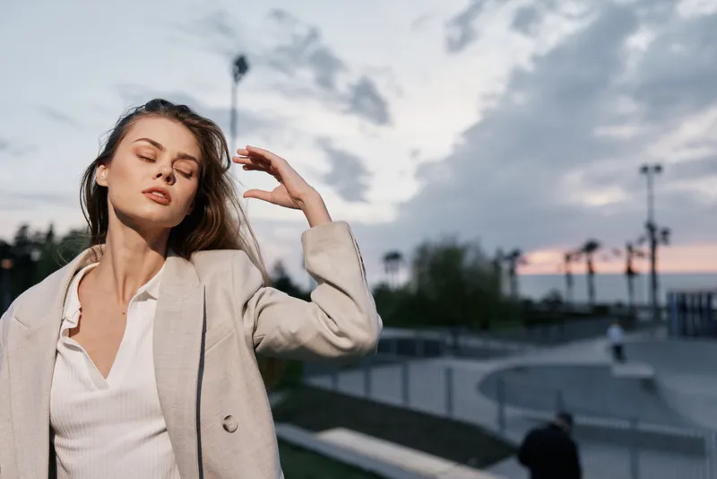 Young woman in beige coat enjoying evening outdoors with eyes closed, serene expression, soft natural light and urban background at sunset key fashion, woman, evening, serene, outdoor, coat, sunset