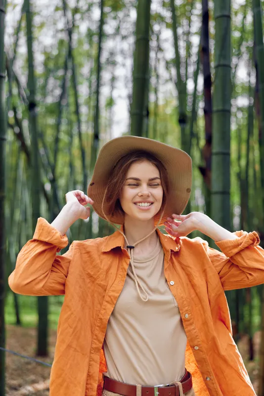 Woman smile hat bamboo forest nature outdoors portrait of a smiling young woman in an orange jacket and widebrim hat, casual fashion, sunlight through green trees and serene relaxed mood.