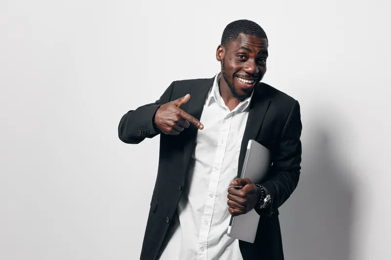 African American man with dark skin wearing black suit and white shirt smiling and pointing at laptop in hand against white background. Business casual style and confident expression.