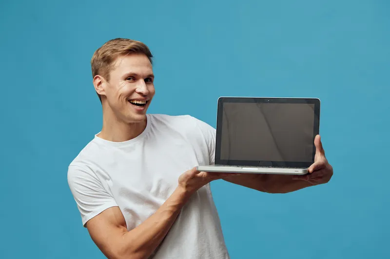 Smiling young man with blond hair holding open laptop computer in front of a plain blue background. Casual attire, joyful expression, presenting technology for online work or digital solutions, studio