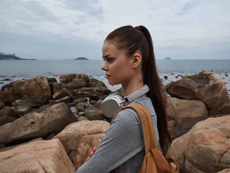 Young woman with long hair wearing headphones by the rocky beach, showcasing a thoughtful expression against a serene ocean backdrop, perfect for lifestyle concepts