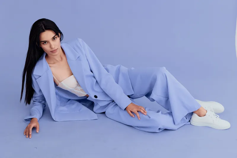 Young woman reclining on purple floor wearing pastel blue suit and white sneakers, dark hair, confident pose, stylish contemporary fashion, minimalist background.