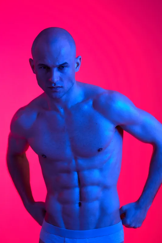 Muscular man posing against a vibrant pink background, showcasing his toned physique. The image highlights bold colors and powerful energy.