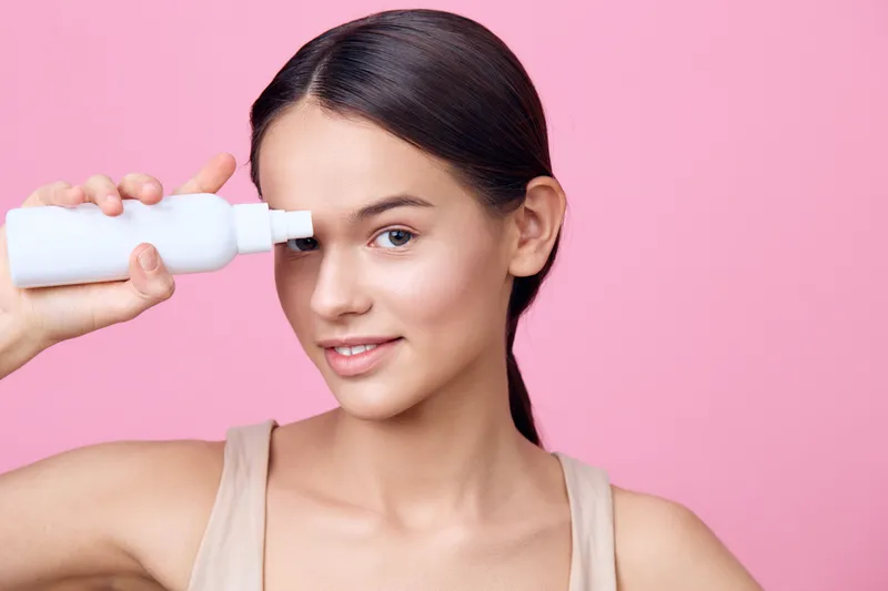Young woman applying skincare product on her face against a soft pink background, promoting healthy skin and beauty routines