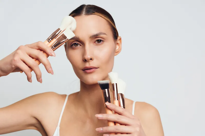 Woman skincare portrait, applying makeup with cosmetic brushes, holding foundation near face, studio beauty shot, calm expression, smooth soft light, clean background