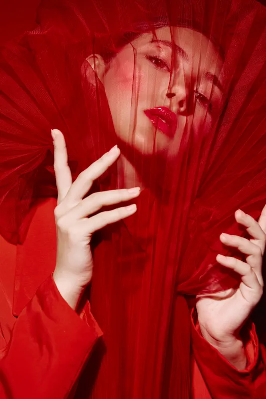 Young woman in creme red makeup and glossy lips posing with a sheer fabric veil. Bold visual in a dramatic red outfit. Concept for beauty, fashion, creative editorial.