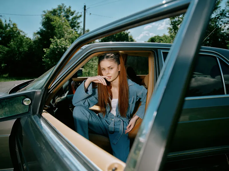 Young woman in trendy casual outfit poses confidently in a luxury car, surrounded by greenery under a bright blue sky, showcasing empowerment and style.