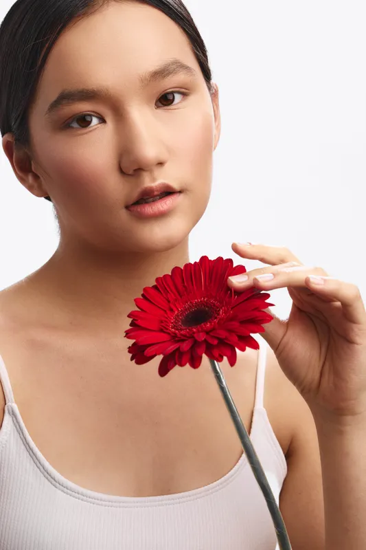 asian woman gazes softly at the camera while holding a red flower near her chest, a minimalist studio setting creating a calm, intimate portrait.