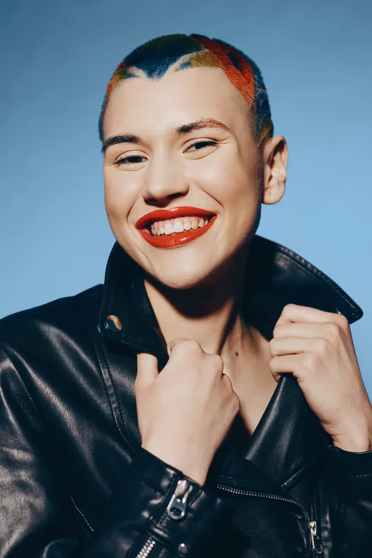 Smiling woman with colorful short hair and bright red lipstick wearing a black leather jacket against a plain blue background, expressing joy and uniqueness.
