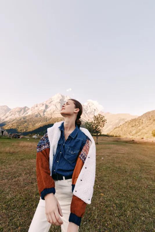 Woman standing in field with mountains in background, wearing patterned sweater and denim shirt, portrait style fashion shot in autumn light, casual model outdoors.