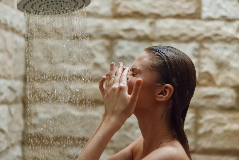 woman showering under rain showerhead, enjoying water droplets, natural stone background, wet hair, skin care, relaxation moment in bathroom