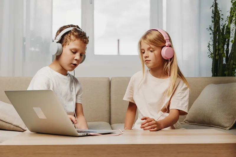 Two children wearing headphones use laptop at home sitting on sofa near window. Focused boy and girl learning or playing with technology in bright living room. Concept of childhood and digital