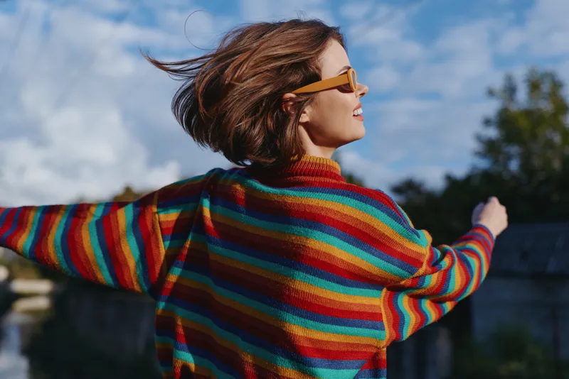 Woman in a rainbow sweater and sunglasses, smiling with arms outstretched outdoors, short hair and casual outfit enjoying a windy urban rooftop scene, joyful travel lifestyle moment.