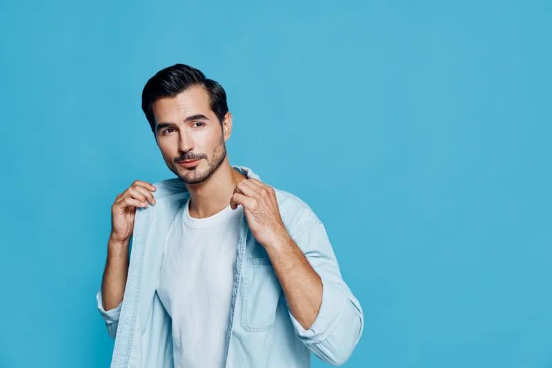 Handsome young man in a light blue shirt poses confidently against a bright blue background, showcasing a fashion-forward style and charisma. People lifestyle concept.