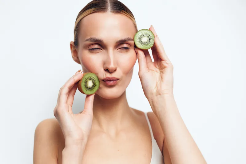 Young woman with clear skin holds two halves of fresh kiwi fruit near her face, posing with closed eyes and puckered lips, isolated on white background for beauty concept.